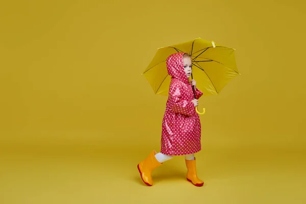 黄色の傘と黄色の背景に赤いレインコートを持つ陽気な子供の女の子 テキストのコピースペース — ストック写真