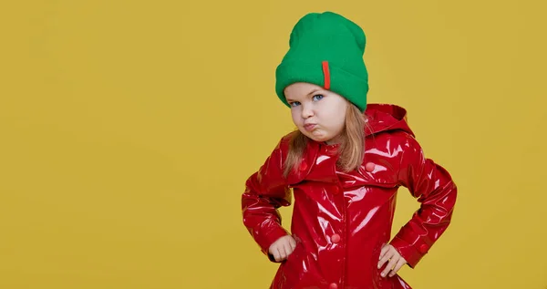赤いレインコートのかわいいスタイリッシュな子供の女の子と色の黄色の背景に緑の帽子 面白い顔 彼女の指を側面に向けテキストのためのスペースをコピー — ストック写真