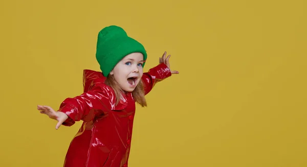 赤いレインコートのかわいいスタイリッシュな子供の女の子と色の黄色の背景に緑の帽子 面白い顔 彼女の指を側面に向けテキストのためのスペースをコピー — ストック写真