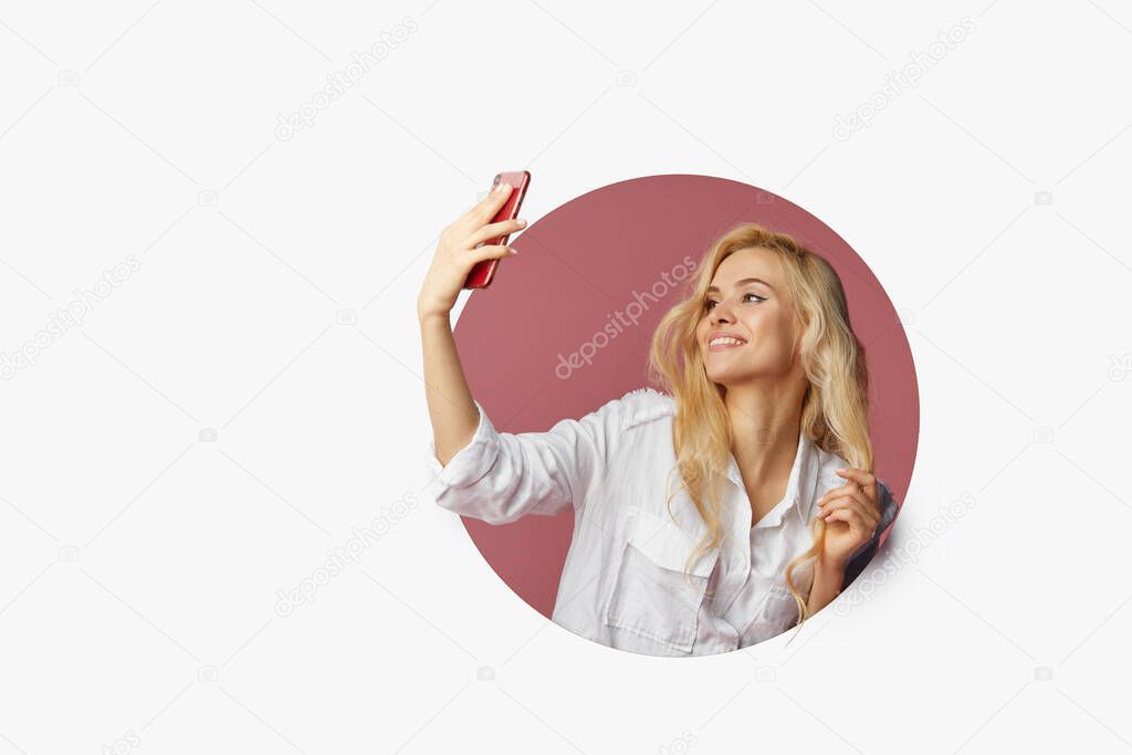 Young beautiful happy woman using a smartphone. Peeks out of a round hole in the wall. White background. Empty space for text.