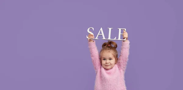 儿童产品的销售 带着购物袋和包装的紫色背景的小女孩的画像 看着摄像机Look Camera 班纳案文的篇幅 — 图库照片