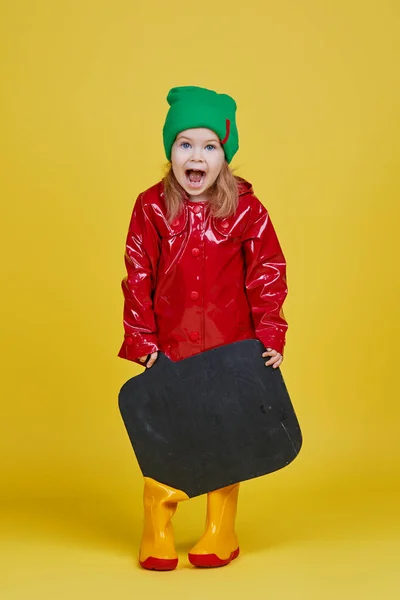 黄色の背景に黒い黒板のバナーを持っている小さな子供の女の子 面白い顔だ テキストのスペースをコピーします 季節の子供製品の広告 — ストック写真