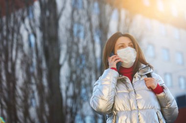 Koruyucu maske takan genç bir kadın güneşli bir günde hastanenin arka planında duruyor. Virüs enfeksiyonu. Covid-19 Coronavirüs Salgını
