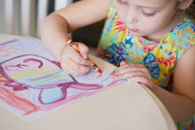 Çocukların yaratıcılığı. Küçük kız evde balmumu kalemlerle resim çiziyor. Küresel karantina dönemi için çevrimiçi uzaklık öğrenme kavramı.