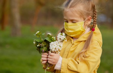 Koruyucu maskeli küçük kız bahçede temiz havada bir buket çiçek tutuyor. Koronavirüs ve küresel karantina pandemik iyileşme, kendini izole etme