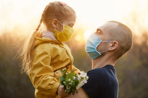 Aile koruması. Genç bir baba bahar bahçesinde küçük kızına sarılıyor. Koronavirüs enfeksiyonundan korunan maskeler.