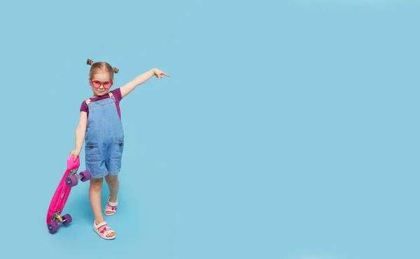穿着鲜艳衣服的可爱的小女孩坐在滑板上 看着蓝色背景的相机 — 图库照片