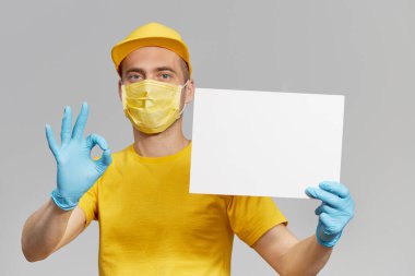 Sarı üniformalı, koruyucu maskeli ve eldivenli kurye paket yemek ve kahve getiriyor. Teslimat servisi karantina altında. Metin için beyaz bir işaret tutuluyor