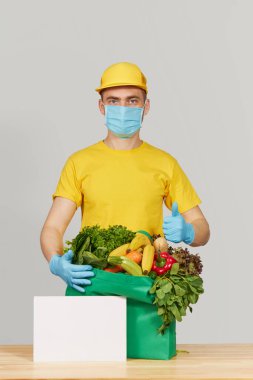 İnternetten alışveriş konsepti. Sarı üniformalı erkek kurye koruyucu maske ve yeni meyve ve sebzelerle dolu bir market kutusuyla eldiven. Koronavirüs karantinası sırasında eve yemek götürme 