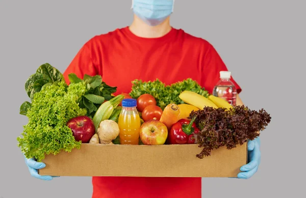 İnternetten alışveriş konsepti. Kırmızı üniformalı erkek kurye, koruyucu maske ve yeni meyve ve sebzelerle dolu bir market kutusuyla eldivenler. Koronavirüs karantinası sırasında eve yemek götürme 