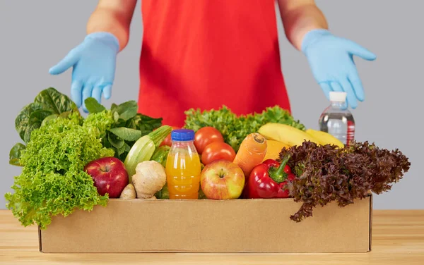 İnternetten alışveriş konsepti. Kırmızı üniformalı erkek kurye, koruyucu maske ve yeni meyve ve sebzelerle dolu bir market kutusuyla eldivenler. Koronavirüs karantinası sırasında eve yemek götürme 