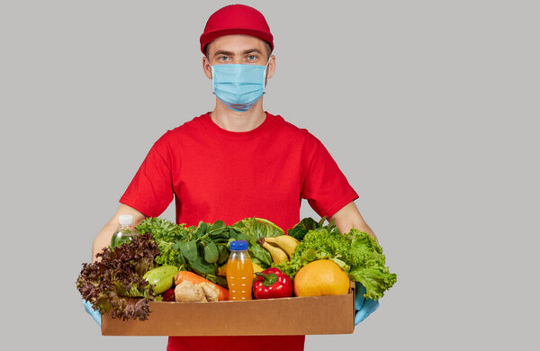 Концепция покупок онлайн. Мужчина курьер в красной форме, защитная маска и перчатки с продуктовой коробкой со свежими фруктами и овощами. Доставка продовольствия на дом во время карантинного коронавируса
 