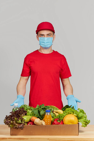 Концепция покупок онлайн. Мужчина курьер в красной форме, защитная маска и перчатки с продуктовой коробкой со свежими фруктами и овощами. Доставка продовольствия на дом во время карантинного коронавируса
 