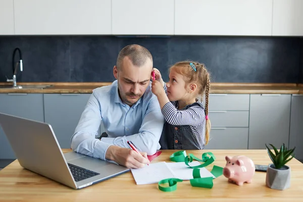 Father Working from home on laptop during quarantine. Little child girl make noise and distracts father from work on the kitchen office