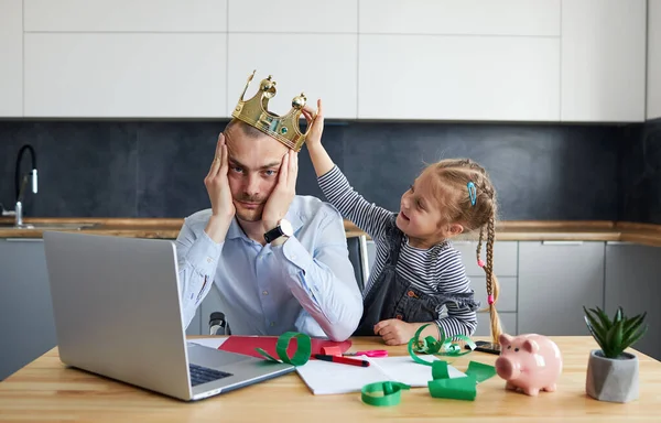 Tired Father Working from home on laptop during quarantine. Little child girl puts a toy crown on dads headon the kitchen office