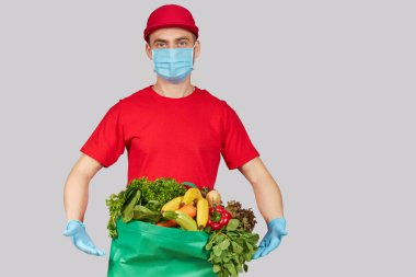 İnternetten alışveriş konsepti. Kırmızı üniformalı erkek kurye, koruyucu maske ve yeni meyve ve sebzelerle dolu bir market kutusuyla eldivenler. Koronavirüs karantinası sırasında eve yemek götürme 