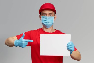 Kırmızı üniformalı, koruyucu maskeli ve eldivenli bir kurye mesaj için beyaz bir işaret tutuyor. Teslimat servisi karantina altında. 