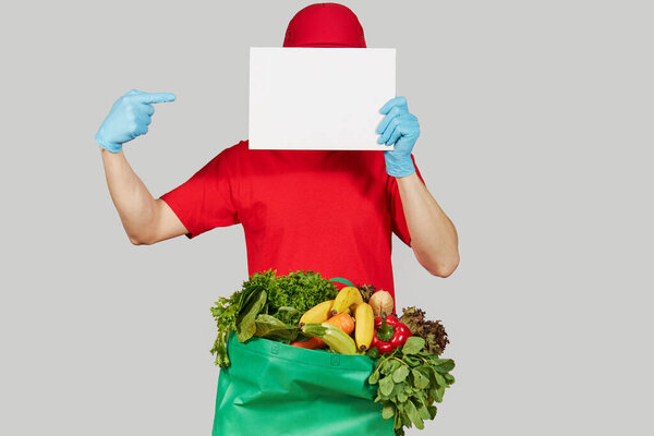 Концепция покупок онлайн. Мужчина-курьер в красной униформе, защитной маске и перчатках с продуктовой коробкой со свежими фруктами и овощами держит белое знамя для текста. Доставка продовольствия на дом во время карантинного коронавируса
 