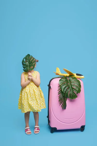 ピンクのスーツケースを着た可愛い女の子が検疫後に熱帯諸国への旅行を待っています ブルースタジオの背景 面白い顔だ — ストック写真