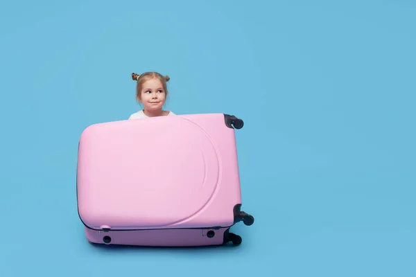 かわいい赤ちゃんの女の子はスーツケースに座って隔離後の夏休みを待っています 側面を指している ブルースタジオの背景 — ストック写真