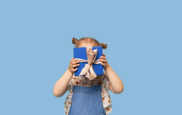 広告赤ちゃん製品 驚くべき興奮した子供の女の子の肖像は青い背景の上に顔の前にギフトボックスを保持しています テキストの空のスペース — ストック写真