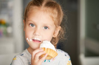 Neşeli, gülümseyen bir kız çocuğu lezzetli bir pasta yiyor ve başparmağını kaldırıyor. Kremalı ağız. Neşeli tasasız bir çocukluk.