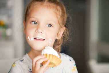 Neşeli, gülümseyen bir kız çocuğu lezzetli bir pasta yiyor ve başparmağını kaldırıyor. Kremalı ağız. Neşeli tasasız bir çocukluk.
