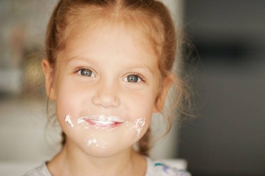 Neşeli, gülümseyen bir kız çocuğu lezzetli bir pasta yiyor ve başparmağını kaldırıyor. Kremalı ağız. Neşeli tasasız bir çocukluk.