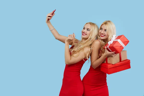 Holidays sales. Happy Women with gift boxes in red dresses on blue background, makes selfie on smartphone. Christmas Gifts.Shopping Mall