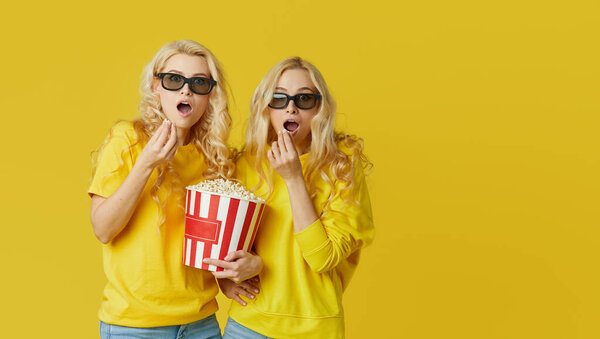 Shocked young models women in 3d glasses eating popcorn, looks scary Movie at the cinema. Isolated on yellow background