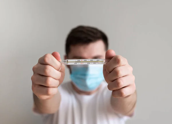 Adam karantinada termometreyle ısıyı ölçüyor. Beyaz adam termometre gösteriyor ve evde karantinada virüs koruması için yüz maskesi ve eldiven takıyor..