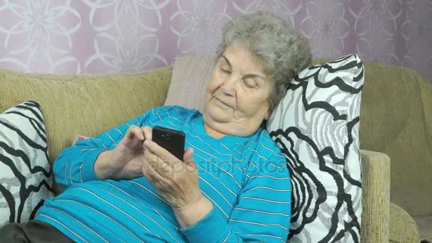 Old woman lying on a beige sofa holds a cellphone — Stock Video