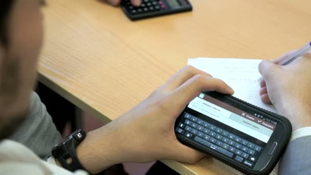 Ο φοιτητής που κάθεται σε ένα γραφείο κατέχει ένα κινητό τηλέφωνο — Αρχείο Βίντεο