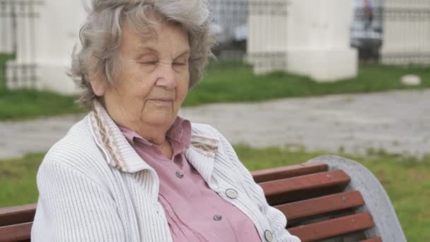 Stará žena s šedými vlasy sedí na lavičce venku — Stock video