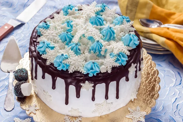 Красивый кремовый торт украшен утечками шоколада, сливками и сахарными снежинками из мастики, на фоне тарелок, чайных ложек и поддонного ножа — стоковое фото