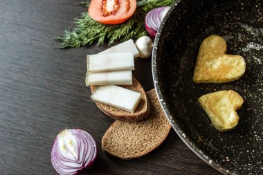 Bir tavada bir kalp şeklinde kızarmış yumurta yakınındaki hangi ekmek dilimleri, domuz yağı (salo), kırmızı soğan, domates, sarımsak, dereotu koyu ahşap açayım.