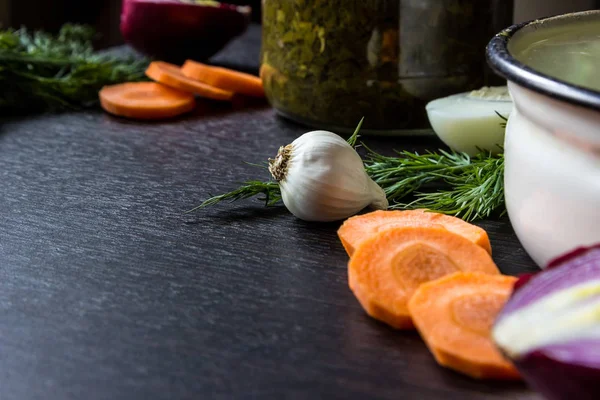 garlic, dill, cut carrot, cut red onion, boiled egg near iron tureen and bowl of sorrel on dark wooden table.