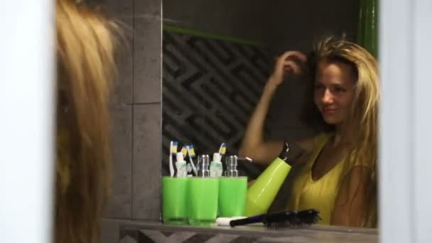 Mujer joven seca el cabello con secador de pelo y sonrisas, reflejo en el espejo . — Vídeos de Stock