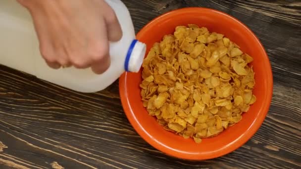 Hand gießt Milch aus einer großen Flasche in die Schüssel voller Cornflakes auf dem Holztisch. Zeitlupe. Ansicht von oben. Nahaufnahme — Stockvideo