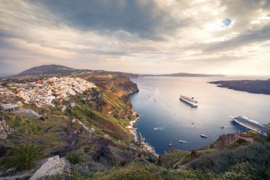 Fira 'yı, Caldera' yı, Santorini yanardağını, günbatımında yolcu gemileriyle muhteşem bir akşam manzarası. Bulutlu dramatik gökyüzü.