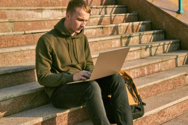 Genç adam dizüstü bilgisayarda çalışıyor kulaklıkla basamaklarda oturuyor ve yanında da bir sırt çantası var.