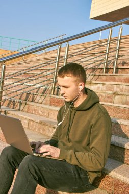 Genç adam dizüstü bilgisayarda çalışıyor kulaklıkla basamaklarda oturuyor ve yanında da bir sırt çantası var.