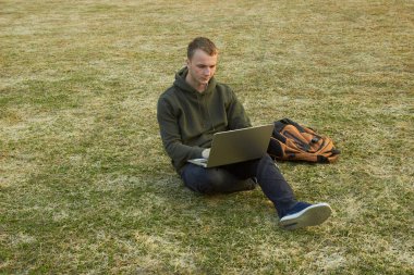Çimlerin üzerinde otururken dizüstü bilgisayarda çalışan yakışıklı bir delikanlının...
