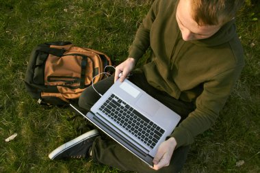 Genç adam laptopta çalışıyor çimlerin üzerinde oturuyor.