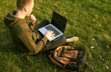 Genç adam laptopta çalışıyor çimlerin üzerinde oturuyor.