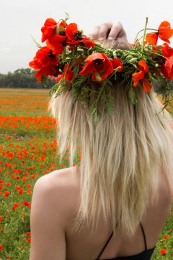 Haşhaş tarlalarının arasında başında gelincik çiçeği çelengi olan güzel, genç bir sarışın kız..