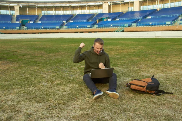 Bilgisayarlı genç adam boş bir futbol sahasında oturuyor.