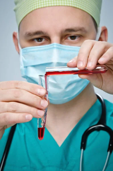 緑の帽子と青いマスクの医者は試験管に試験管から赤い液体を注ぐ. — ストック写真