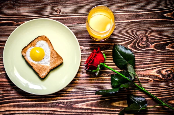 Breakfast consists of eggs in the form of heart, toast and juice. Next to the breakfast is a red rose. — Stock Photo, Image