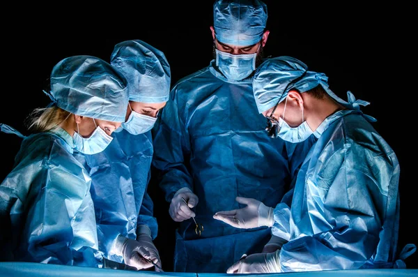 El asistente envía el instrumento al médico durante la operación. Los médicos usan trajes quirúrgicos azules y gorras quirúrgicas, en sus caras tienen máscaras médicas. . —  Fotos de Stock